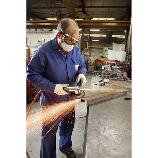 Facom V_472F angle grinder in use, producing sparks on metal, workshop setting, operator in blue coveralls and gloves.